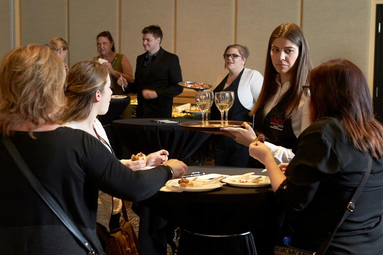 A group gathers and eats while a student serves wine from a tray