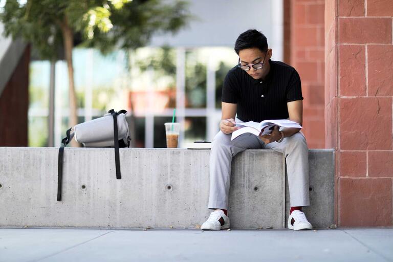 A student sits on a cement bench and reads a book next to a backpack and iced coffee.