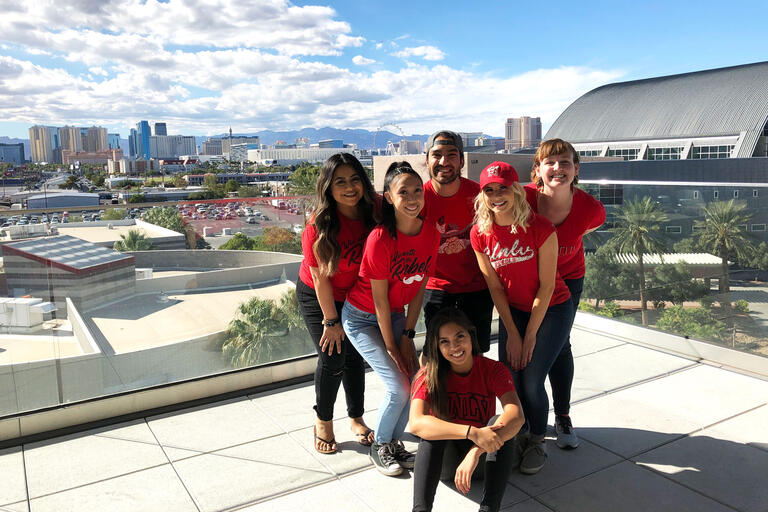 A group of six students wearing red tshirts stand on an outdoor patio with a view of Las Vegas in the background.