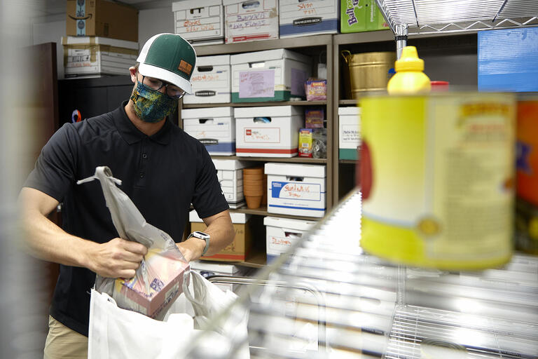 man wearing face mask working putting grocery items in bag in storeroom