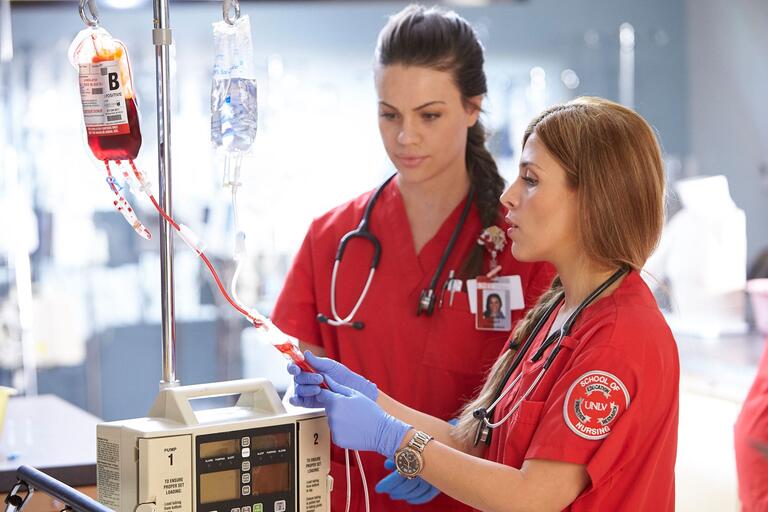 Two nursing students in red scrubs work with an IV in the SIM lab