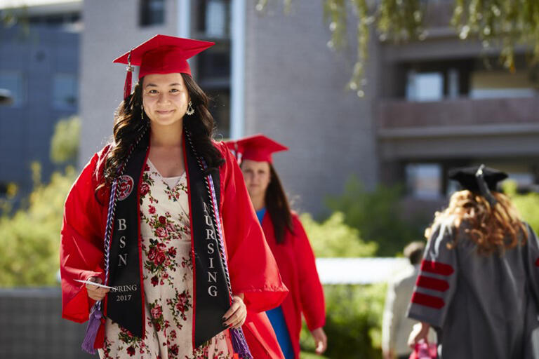 Young woman wearing a red commencement cap and gown over a floral dress. She is also wearing a U-N-L-V Nursing sash
