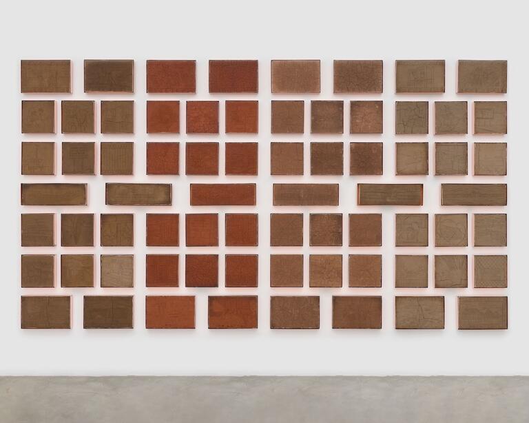 A mass of earth-brown squares and rectangles is arranged in seven rows on a wall. They form a block of modulated hues—the ones on the right are a lighter shade of brown, the one on the left are darker, and the ones in between become reddish—with a grid of white gaps running between their edges. Line drawings of animals, landscapes, and urban infrastructure have been incised into the brown surfaces. Some are cracked like dry earth.
