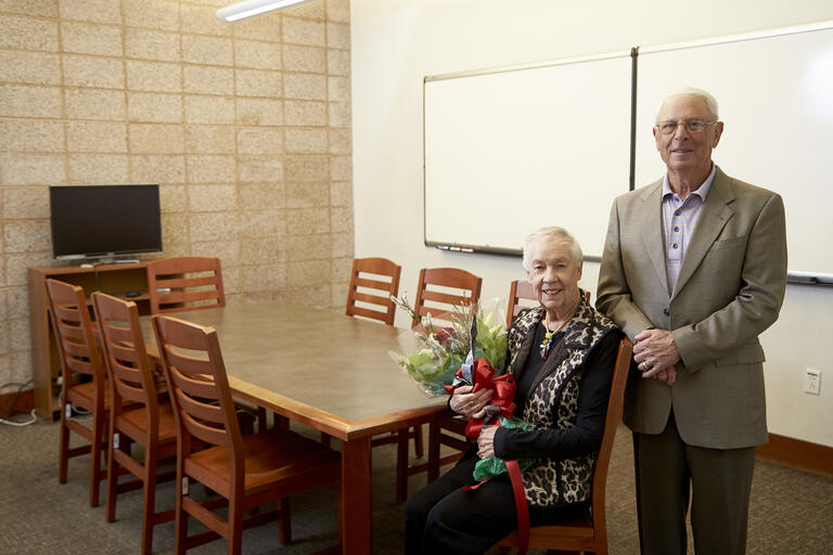 older man and woman, who is holding flowers, pose by table