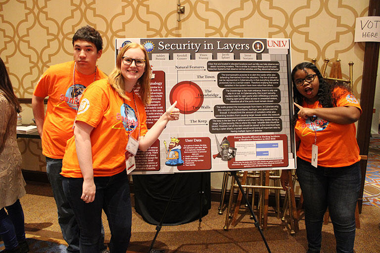 Three students pointing to a poster about Security in Layers