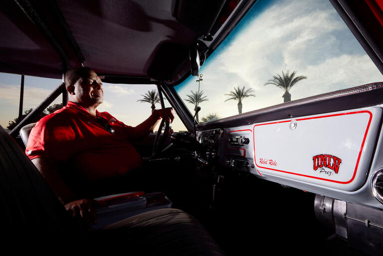 man behind the wheel of a classic car with palm trees and sky in distance