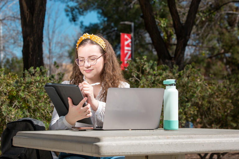 Student using a tablet and laptop outside in front of a UNLV banner.