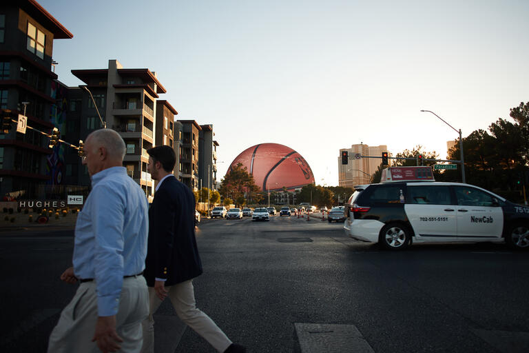 Two men crossing the street in Las Vegas with The Sphere at the Venetian in the background. The Sphere is lit up as a basketball.