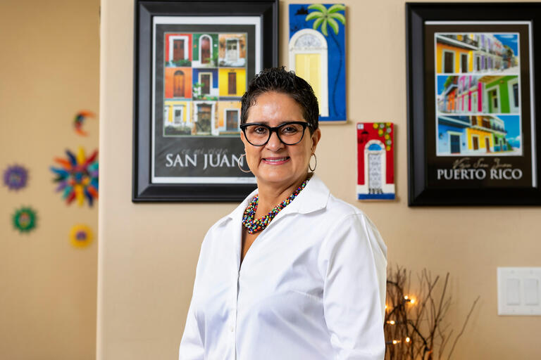 A professional woman in front of two framed photos, one of San Juan and one of Puerto Rico