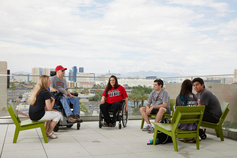 Student candid group photo on campus. Two of the students are using mobility aids and the rest of the students are sitting in green chairs. They are on the plaza overlooking The Strip.