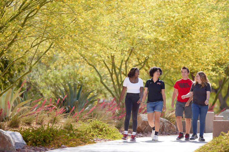 four college students walking on path with green trees and plants