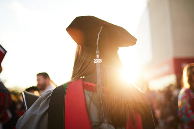 A graduate student whose back is turned to the camera is wearing a cap with a 2023 tassel and walking into a crowd of students