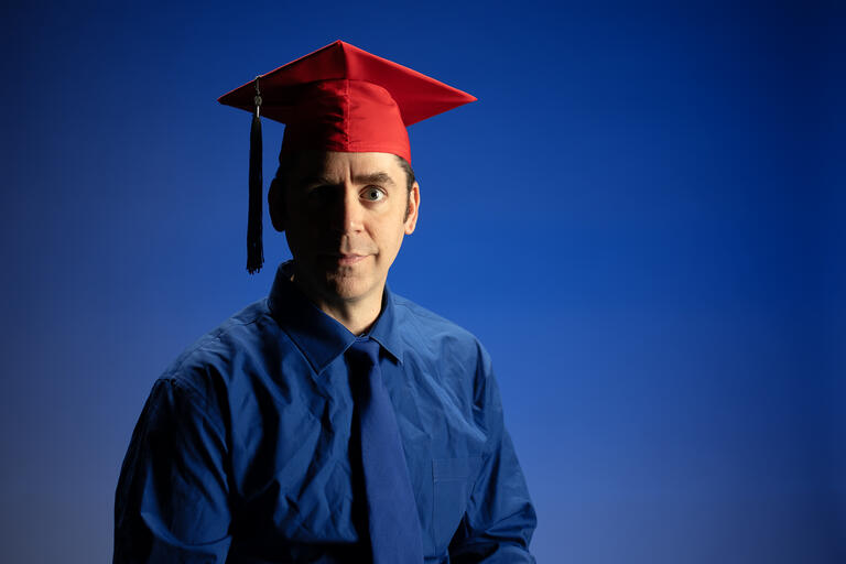 A portrait of Kirk Massey in blue clothes in front of a blue backdrop. He is wearing a red graduation cap.
