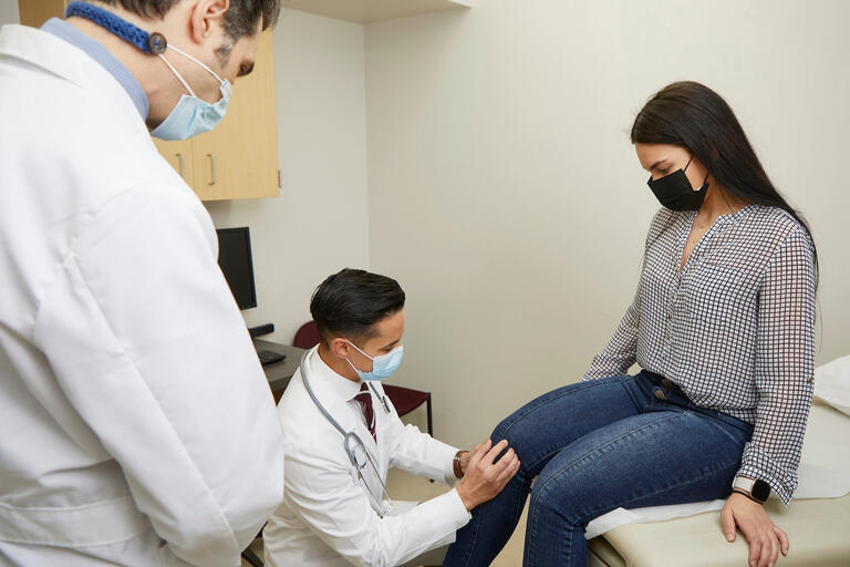 Two doctors in a room with a patient. One doctor assesses the patient's knee while the other observes.