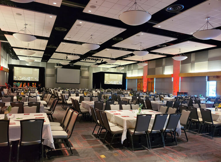 A ballroom with sets of square shaped tables and 12 chairs and screens on stage