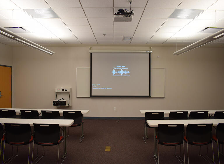 A room with rows of tables and chairs, a whiteboard, and a projector