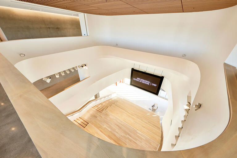 Interior view of a modern, multi-level atrium featuring curved white walls, wooden accents, and a wide wooden staircase.