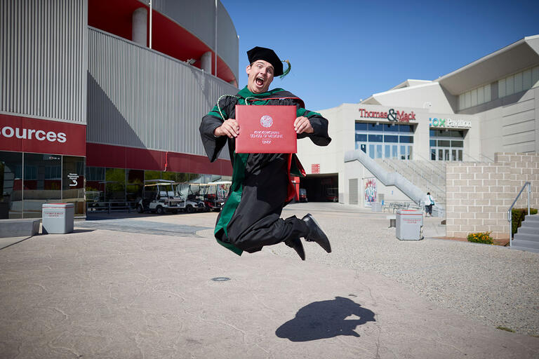 A graduate in cap and gown jumps in excitement, holding their diploma.
