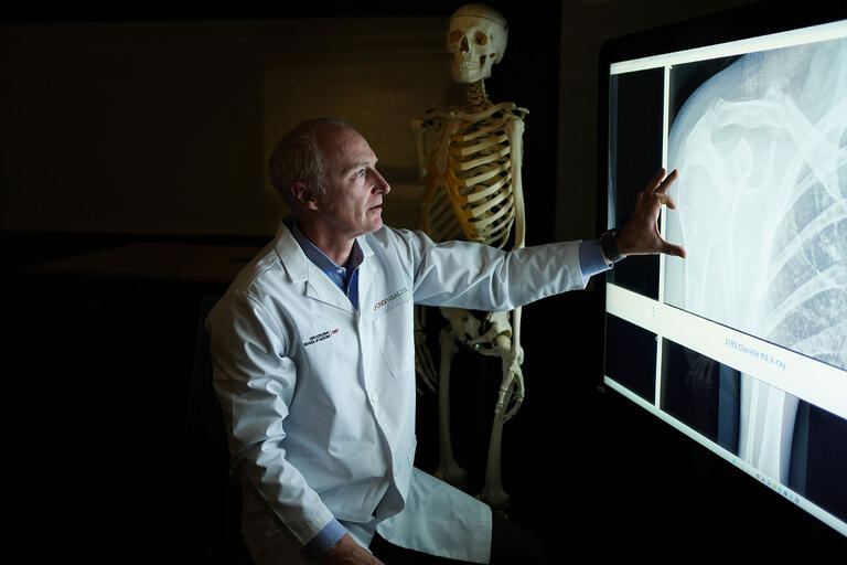 A male doctor in a white lab coat examines a shoulder X-ray on a large digital screen.