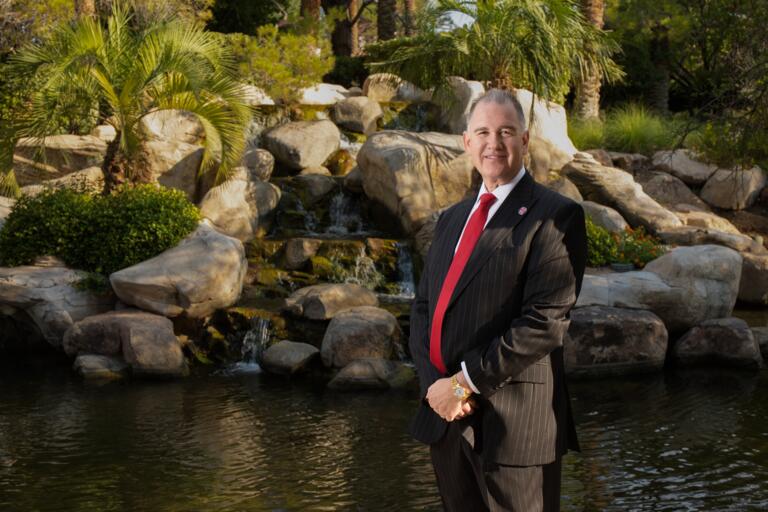 Older white man with receding gray hair wearing a black pinstripe suit, white collared shirt, red tie and gold watch on his left wrist poses with his hands crossed in front of him with small lake, waterfall, large boulders and palm trees in the background