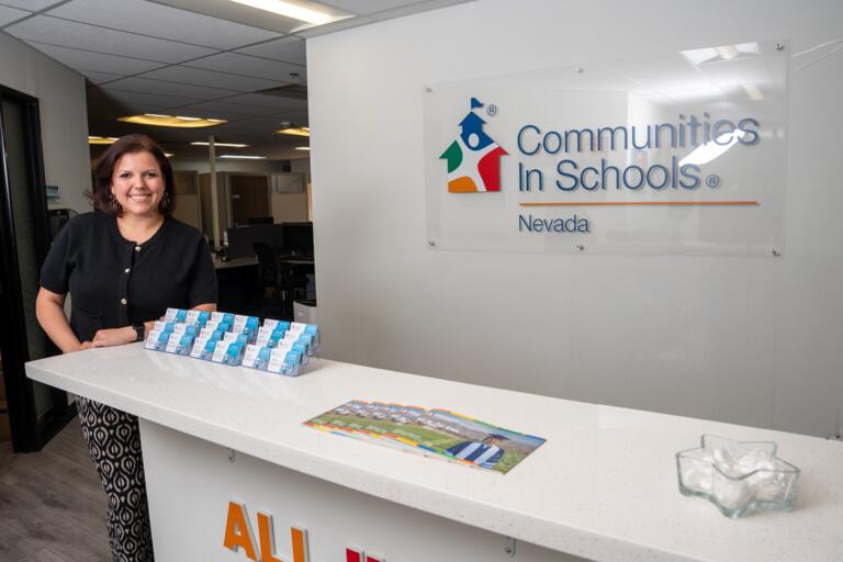 Latin woman in a black top with gold buttons smiles as she leans against the front-desk counter at Communities in Schools Nevada