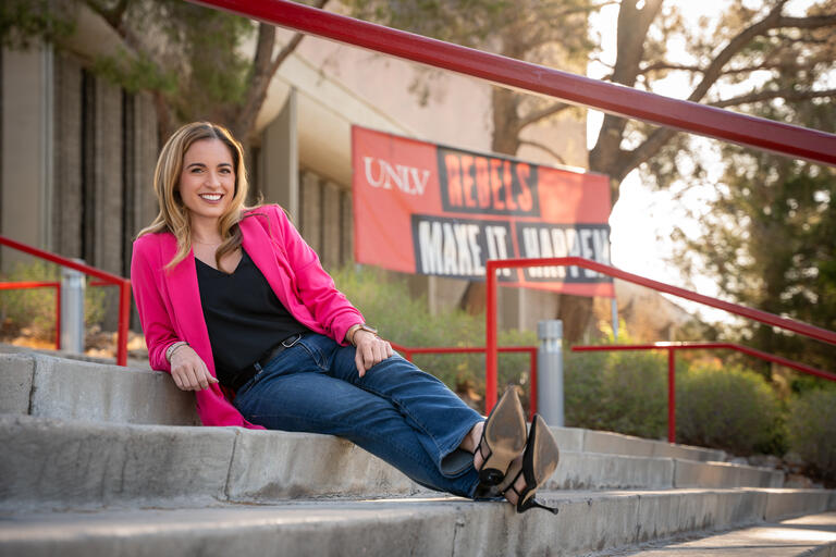 Blonde woman wearing blue jeans and black shirt covered by a pink lightweight jacket extends her legs as she relaxes on steps outside on UNLV's campus
