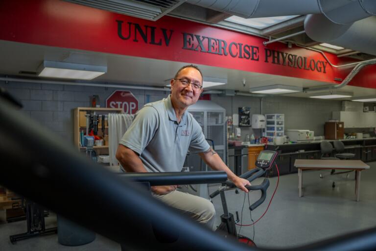 Older white man with receding brown hair wearing tan pants, a gray UNLV polo shirt and eyeglasses sits on an exercise bike in the UNLV Exercise Physiology lab