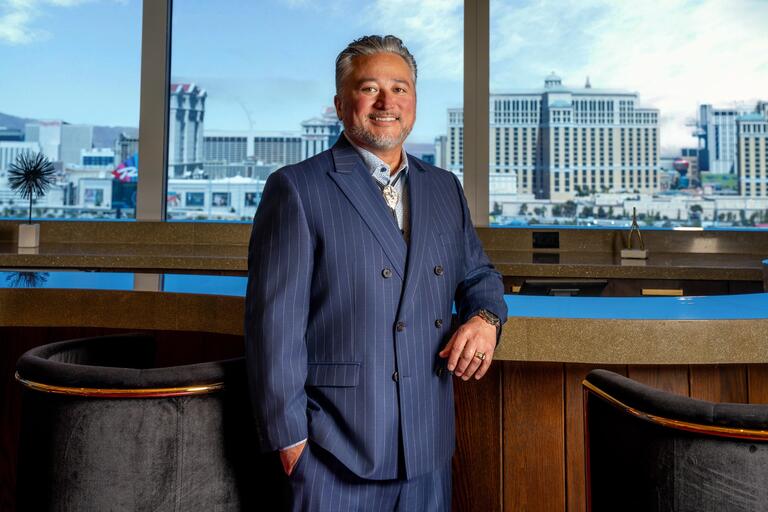 Man in a dark blue pinstripe suit with black and gray hair and beard poses in a hotel bar with his left arm resting on the bar and the Bellagio hotel in the background