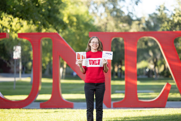 Amelia Davis holds holiday cards she designed in front of UNLV letters sculpture