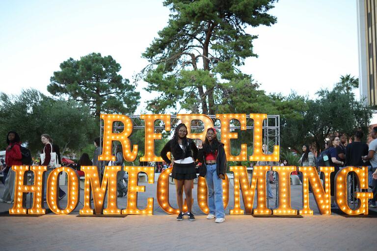 Two students stand in front of a large gold Rebel Homecoming sign lit up with lights
