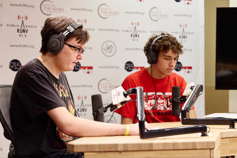 2 students talk into radio mics. Behind, the wall reads KUNV Radio.