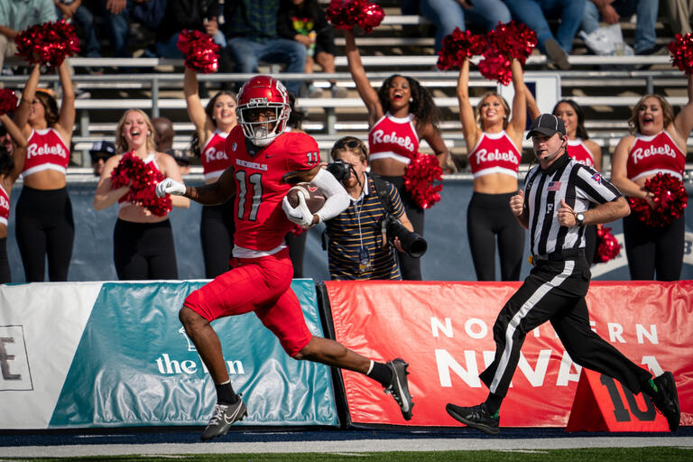 A football player runs across the field, passing a referee. UNLV cheerleaders perform in the background.