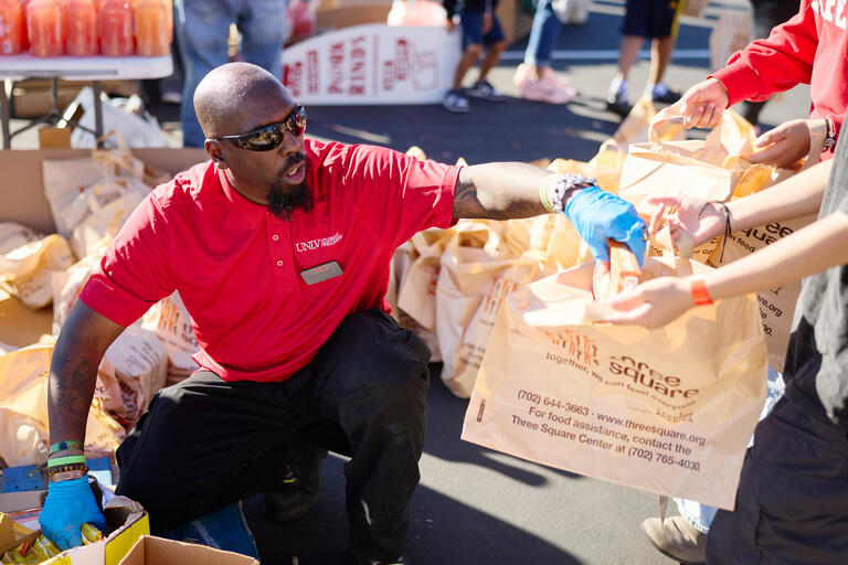 A man wearing a red UNLV polo places food into a plastic bag bearing a Three Square logo