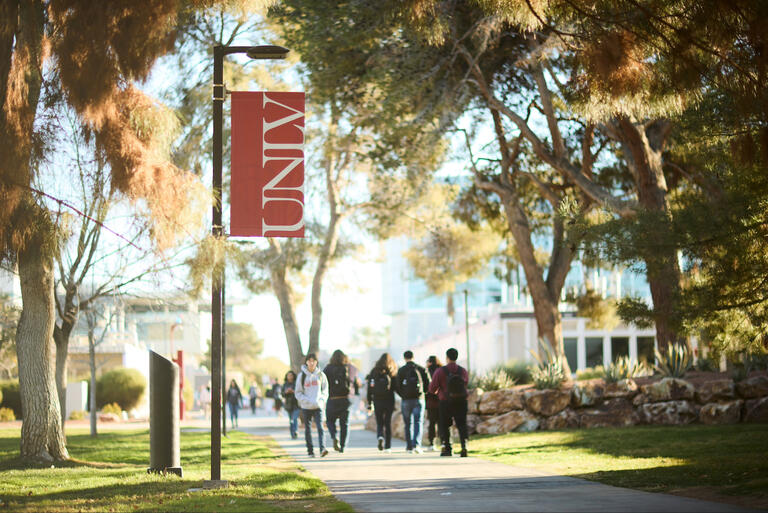 Students walking on campus with a UNLV banner pole in the foreground.