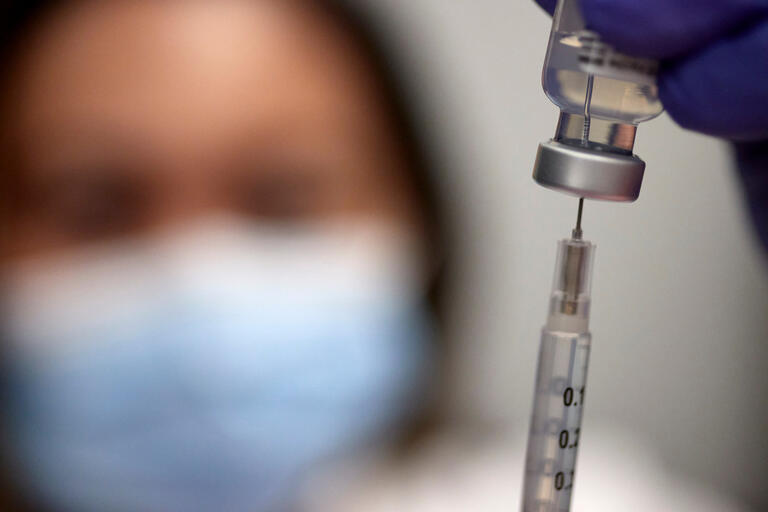 A masked medical worker inserts a syringe into a vial containing COVID vaccine solution