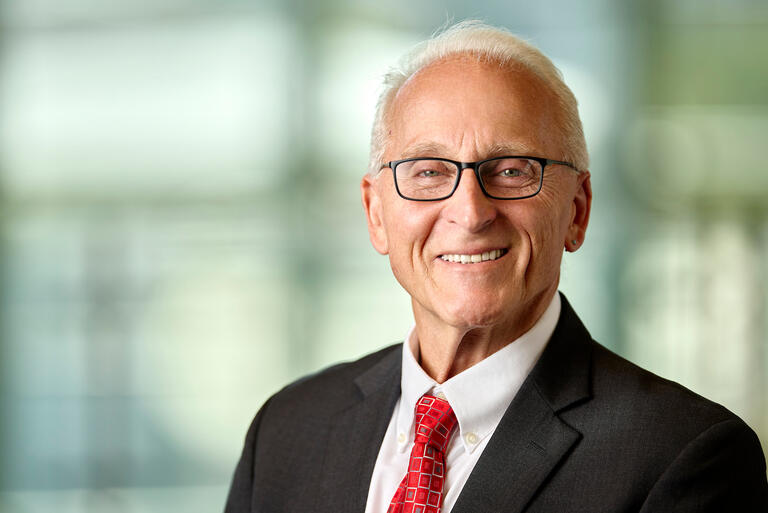 Older man with short gray hair smiles as he poses against a blurred background while wearing a white collared shirt, black suit jacket, red checkered tie and eyeglasses
