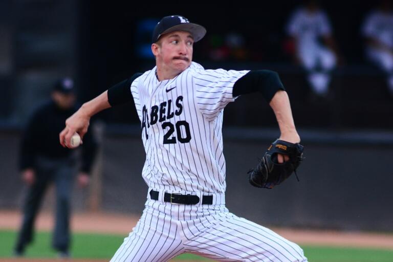White male baseball pitcher in a black and white pinstripe Rebels uniform with the No. 20, black stirrup socks and a black baseball cap grips a baseball in his right hand, reaches behind himself and prepares to deliver a pitch