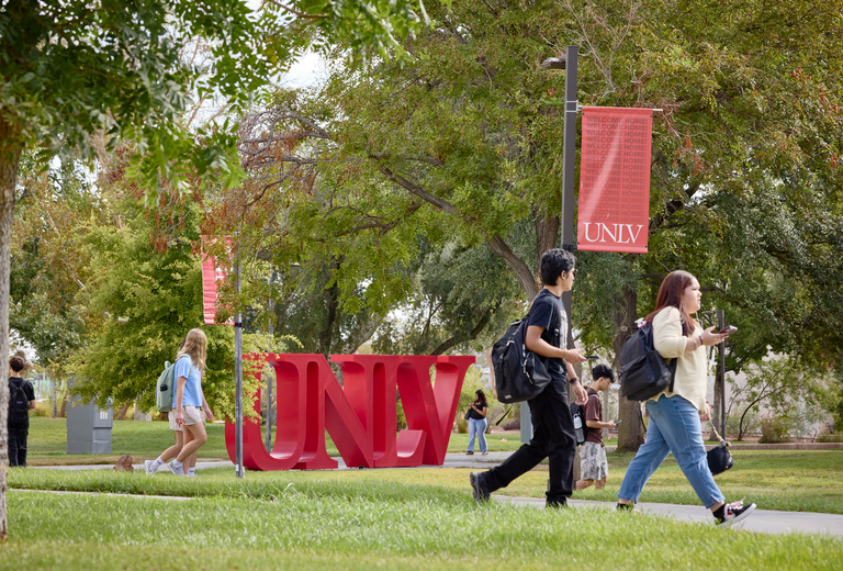 Students walk together down a campus sidewalk. A large, red UNLV sign is visible in the background
