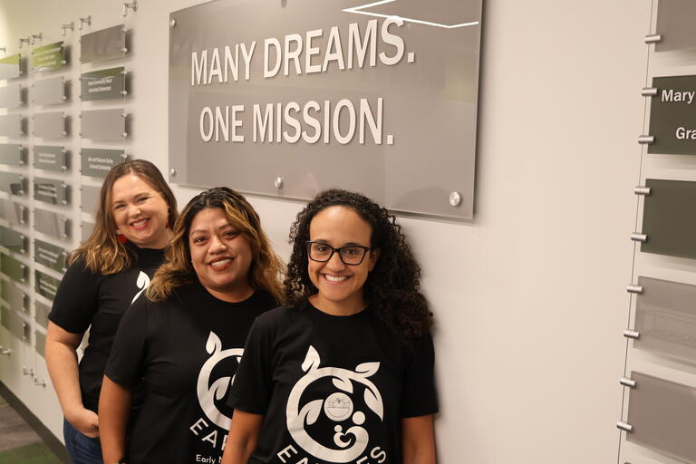 three women under a sign that reads "many dreams. one mission."