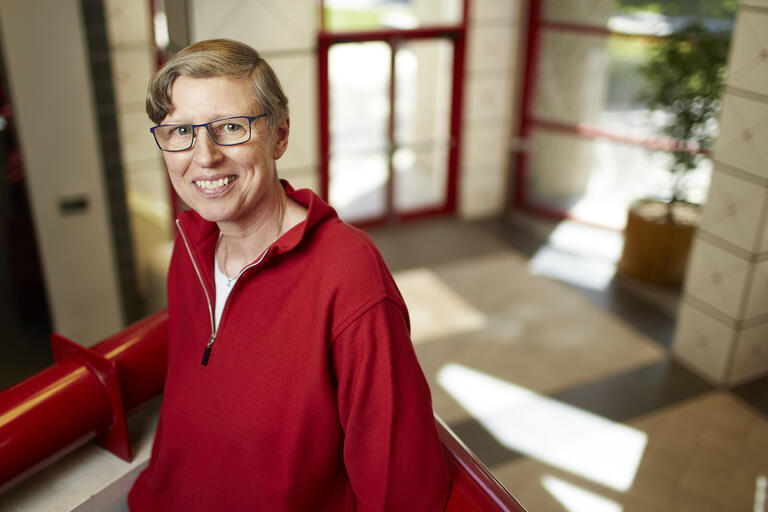 Gabriele Wulf is pictured in a red sweater standing on a landing in the lobby of the Bigelow Health Sciences on UNLV's campus