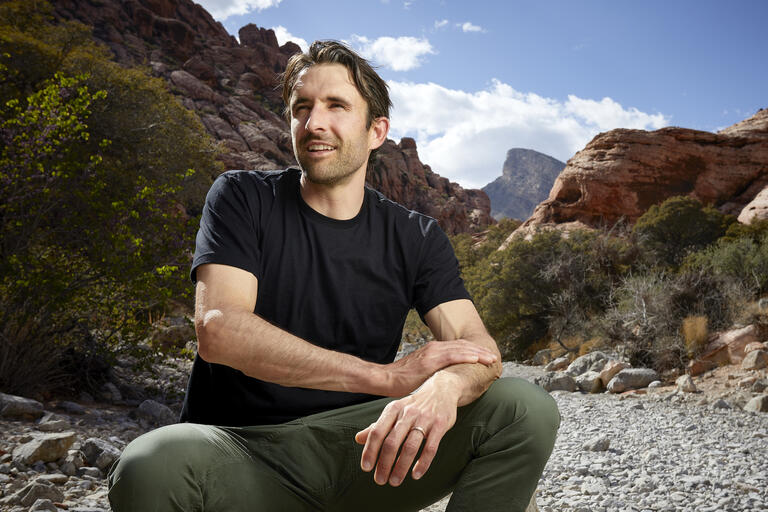 UNLV journalism professor pictured sitting on a rock in Calico Basin with mountains and blue sky in the background