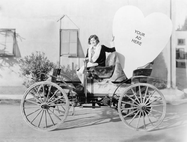 1910s woman sitting in vintage car with large cardboard heart that reads: Your Ad Here