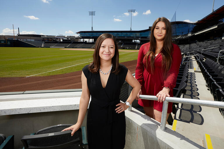 Two women in business attire standing in the box seats of a new baseball stadium.