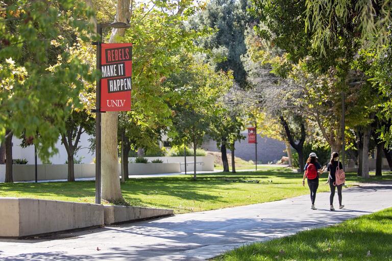 UNLV walkway with students walking on sidewalk, passing sign that says Rebels Make It Happen