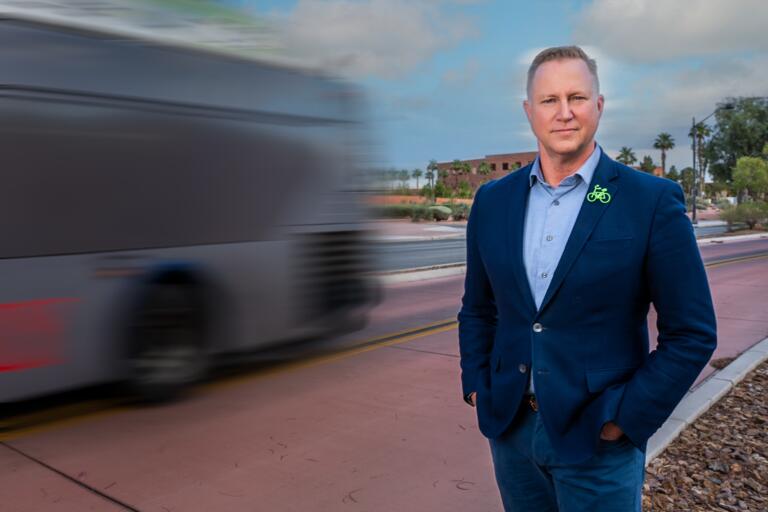 White man in a navy blue blazer, light blue button-down shirt and blue jeans poses with his hands in his pockets in front of a blurred image of a city bus