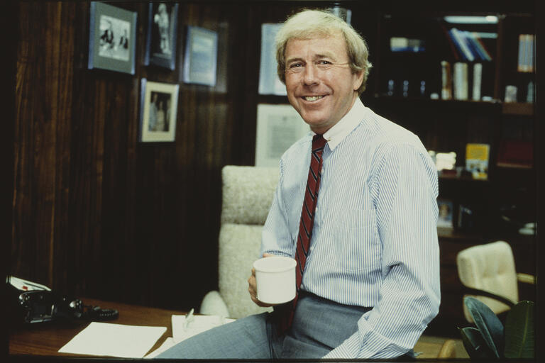 archive photo of man in tie holding coffee cup and leaning on office desk