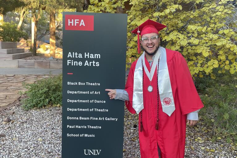 UNLV grad in cap and gown pointing at a Department of Art sign on campus