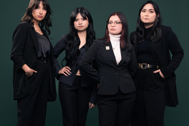 Four women in suits posing for the camera against a dark green background