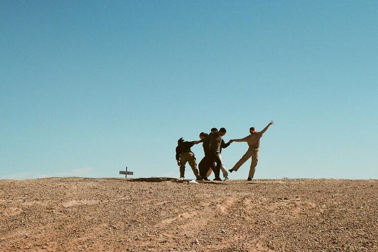 A desert landscape with a handful of dancers in beige clothing in mid pose.