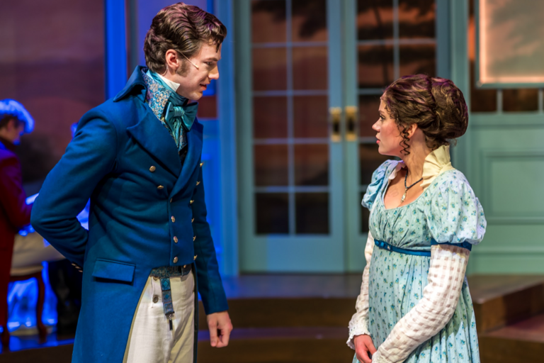 Two actors on stage for a play. Man in a navy blue suit, woman in a baby blue dress. They are speaking to each other.